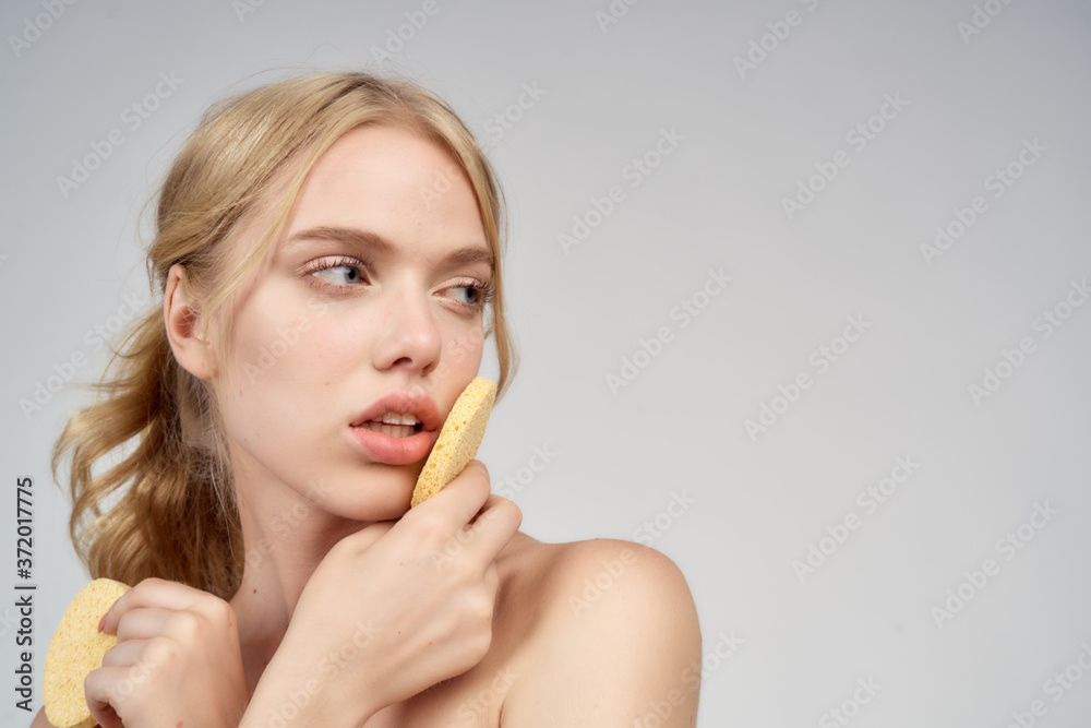 A blonde with bare shoulders holds cotton pads near her face skin care light background