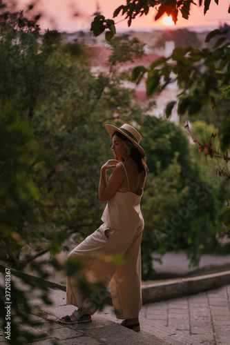 Young beautiful woman wearing straw hat and linen beige suit posing on a city streetat sunset.