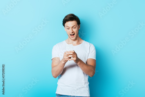 Excited man in white t-shirt using smartphone on blue background