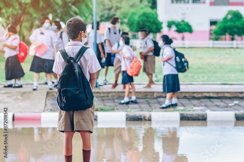 Male elementary school student wear face mask to prevent the Coronavirus(Covid-19) wait for her parents to pick her up to return home after school and the rain just stop in front of the school gate