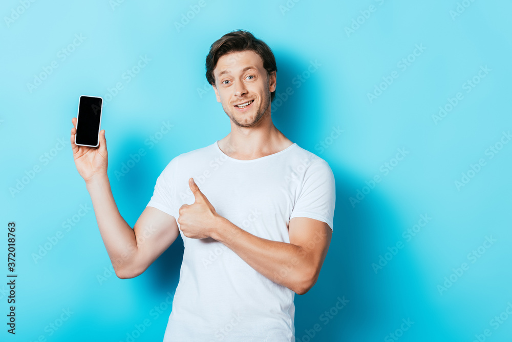 Young man showing thumb up and smartphone with blank screen on blue background Young man showing thumb up and smartphone with blank screen on blue background