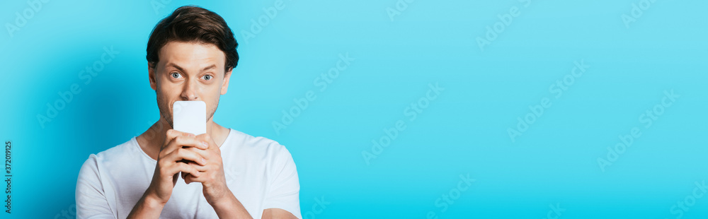 Panoramic shot of young man looking at camera while holding smartphone near mouth on blue background Panoramic shot of young man looking at camera while holding smartphone near mouth on blue background