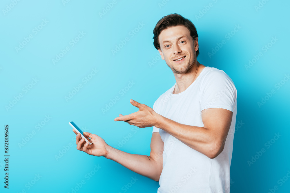 Young man in white t-shirt pointing with hand at smartphone on blue background Young man in white t-shirt pointing with hand at smartphone on blue background