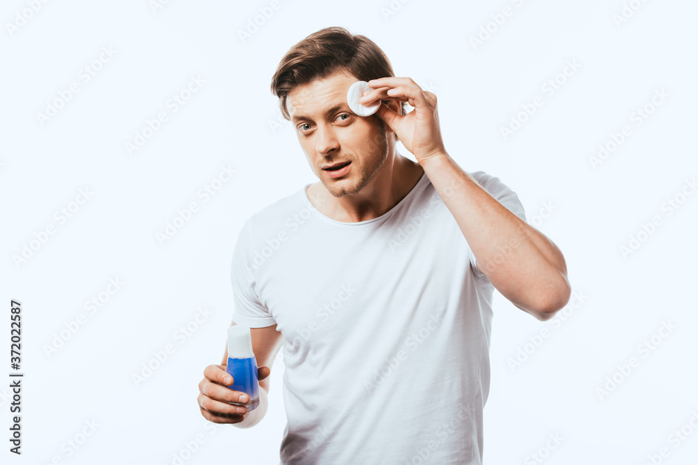 Young man looking at camera while applying lotion with cotton pad isolated on white Young man looking at camera while applying lotion with cotton pad isolated on white