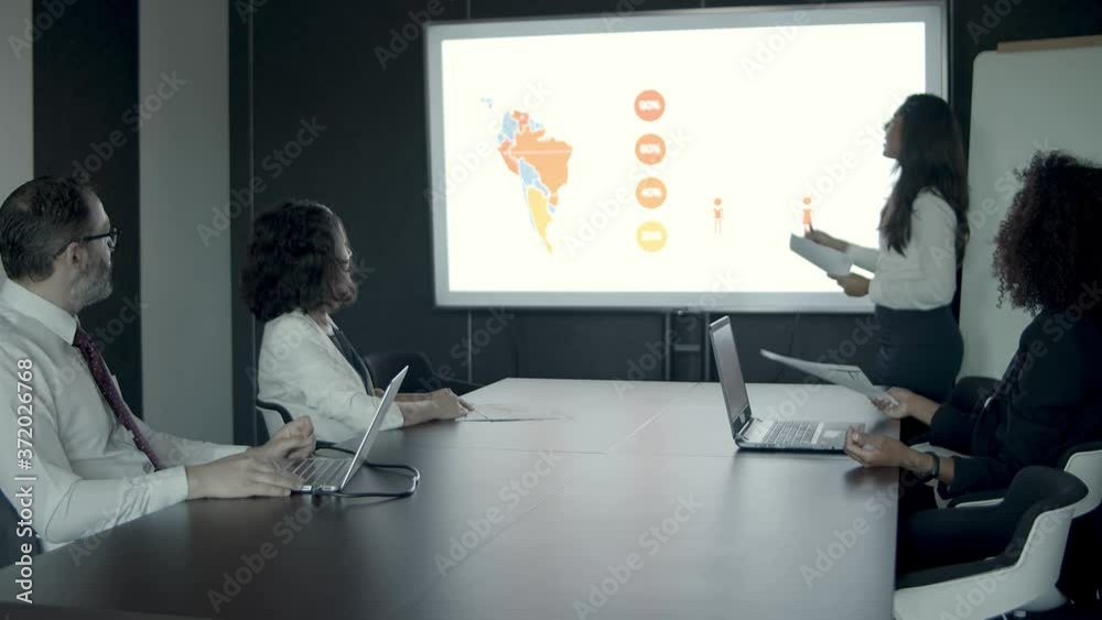 Female office worker explaining data and pointing to charts with maps. Professional businesspeople carefully listening report and sitting in conference room. Business team and presentation concept Female office worker explaining data and pointing to charts with maps. Professional businesspeople carefully listening report and sitting in conference room. Business team and presentation concept