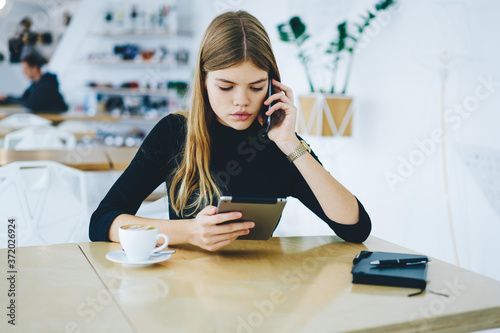 Beautiful manager having conversation via smartphone while watching video on modern touch pad sitting in coworking space with coffee.Attractive female talking on cellular while browsing digital tablet