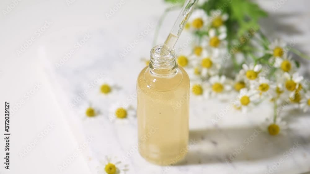 Essential of camomile flowers oil in a dropper glass bottle. Drops falling from pipette into an antiaging moisturizing product. Marble table Essential of camomile flowers oil in a dropper glass bottle. Drops falling from pipette into an antiaging moisturizing product. Marble table