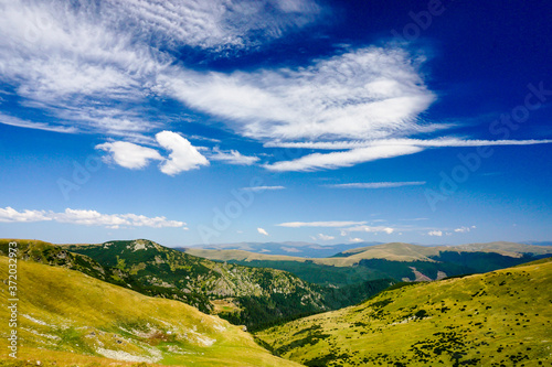 mountain landscape, green hills with grass and trees in the distance, blue sky with few white clouds, near Translapina road in Romania
