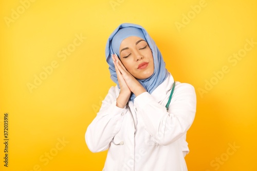 Young beautiful Arab doctor woman wearing medical uniform standing over yellow background sleeping tired dreaming and posing with hands together while smiling with closed eyes.