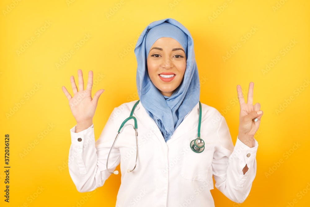 Φωτογραφία young beautiful Arab doctor woman wearing medical uniform standing over yellow background showing and pointing up with fingers number seven while smiling confident and happy