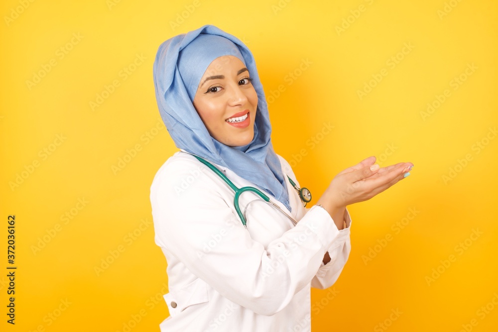 young beautiful Arab doctor woman wearing medical uniform standing over yellow background pointing aside with hands open palms showing copy space, presenting advertisement smiling excited happy young beautiful Arab doctor woman wearing medical uniform standing over yellow background pointing aside with hands open palms showing copy space, presenting advertisement smiling excited happy