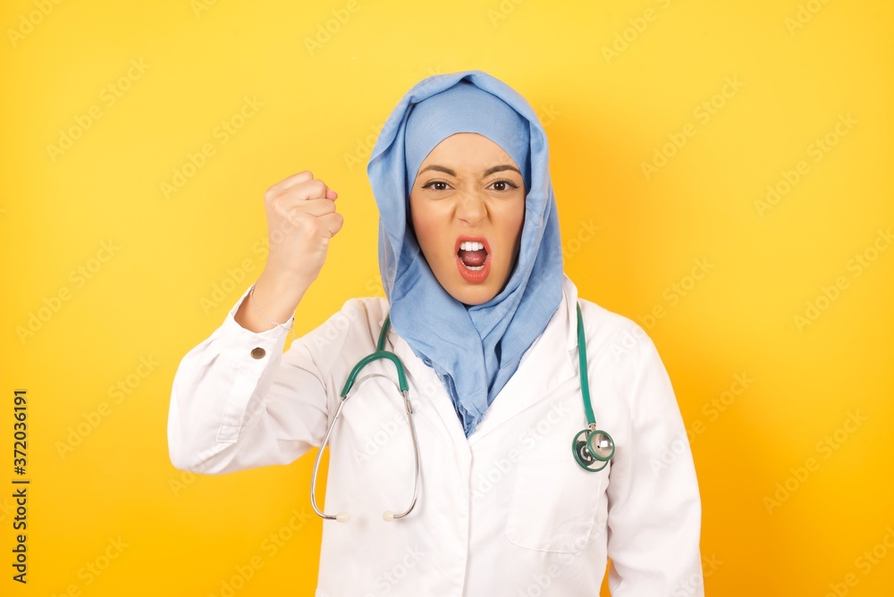 Φωτογραφία young beautiful Arab doctor woman wearing medical uniform standing over yellow wall, being angry and mad raising fist frustrated and furious while shouting with anger