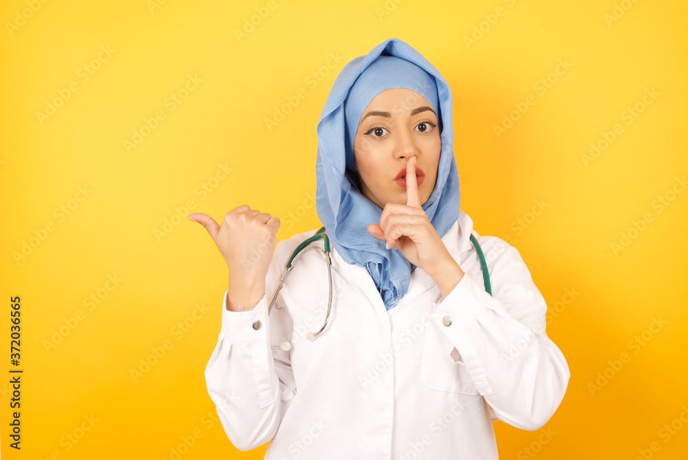 Φωτογραφία Young arab doctor man wearing medical uniform standing over yellow background , asking to be quiet with finger on lips pointing with hand to the side