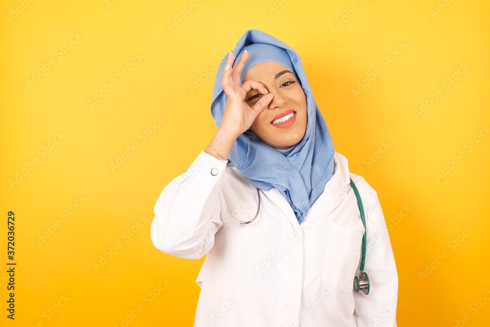 Young arab doctor woman wearing medical uniform standing over yellow background doing ok gesture with hand smiling, eye looking through fingers with happy face. Young arab doctor woman wearing medical uniform standing over yellow background doing ok gesture with hand smiling, eye looking through fingers with happy face.