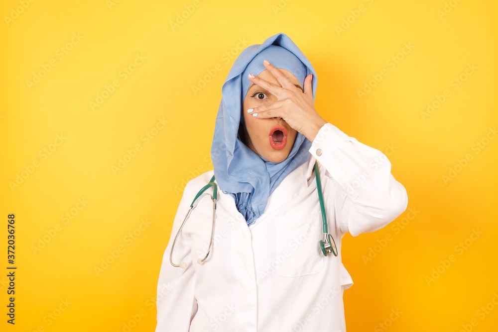 Young arab doctor woman wearing medical uniform standing over yellow background peeking in shock covering face and eyes with hand, looking through fingers with embarrassed expression. Young arab doctor woman wearing medical uniform standing over yellow background peeking in shock covering face and eyes with hand, looking through fingers with embarrassed expression.