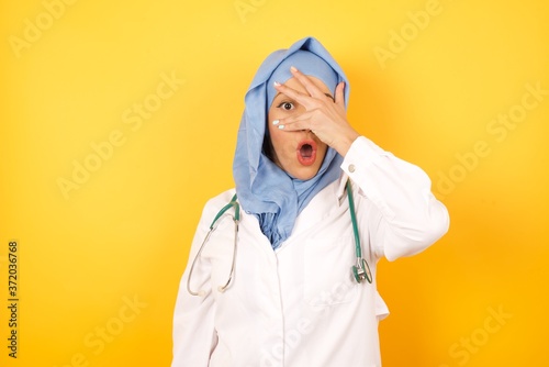 Young arab doctor woman wearing medical uniform standing over yellow background  peeking in shock covering face and eyes with hand, looking through fingers with embarrassed expression. © Roquillo