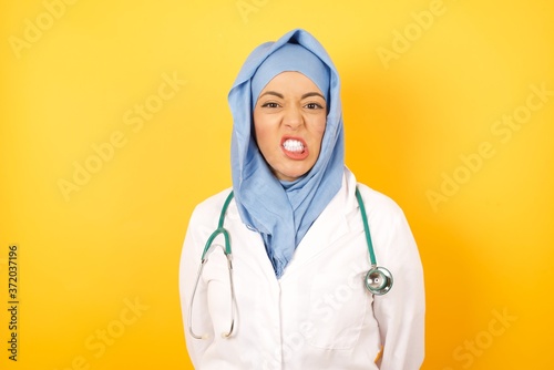 Portrait of angry Young arab doctor woman wearing medical uniform standing over yellow, keeps teeth clenched, frowns face in dissatisfaction, irritated because of much duties. , sees miserable weather