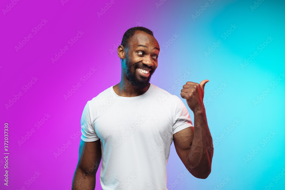Pointing, showing. African-american young man's portrait on gradient studio background in neon. Beautiful male model in casual style, white shirt. Concept of human emotions, facial expression, sales Pointing, showing. African-american young man's portrait on gradient studio background in neon. Beautiful male model in casual style, white shirt. Concept of human emotions, facial expression, sales