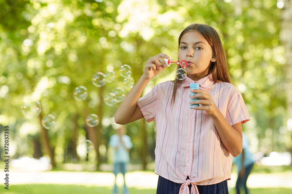 Waist up portrait of teenage girl blowing bubbles and looking at camera while standing in green park outdoors, copy space