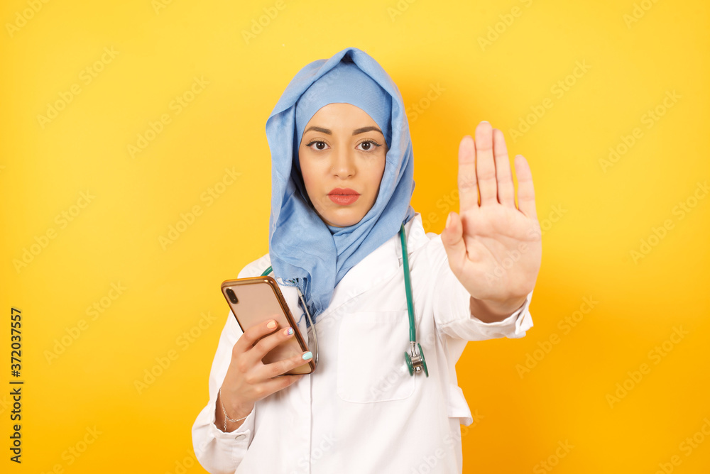 Portrait of beautiful muslim doctor woman wearing medical uniform  using and texting with smartphone over isolated background with open hand doing stop sign with serious and confident expression, 
