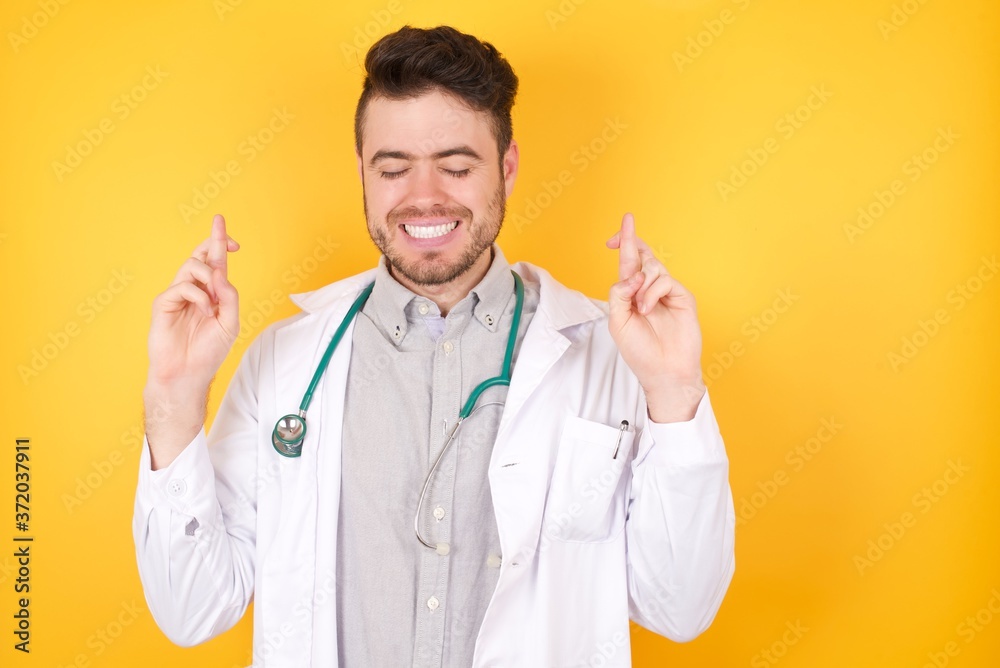 Photo of positive Caucasian doctor man has big hope, crosses fingers, wears medical uniform, believes in good fortune, smiles broadly, isolated over yellow background. People and wish concept. Photo of positive Caucasian doctor man has big hope, crosses fingers, wears medical uniform, believes in good fortune, smiles broadly, isolated over yellow background. People and wish concept.