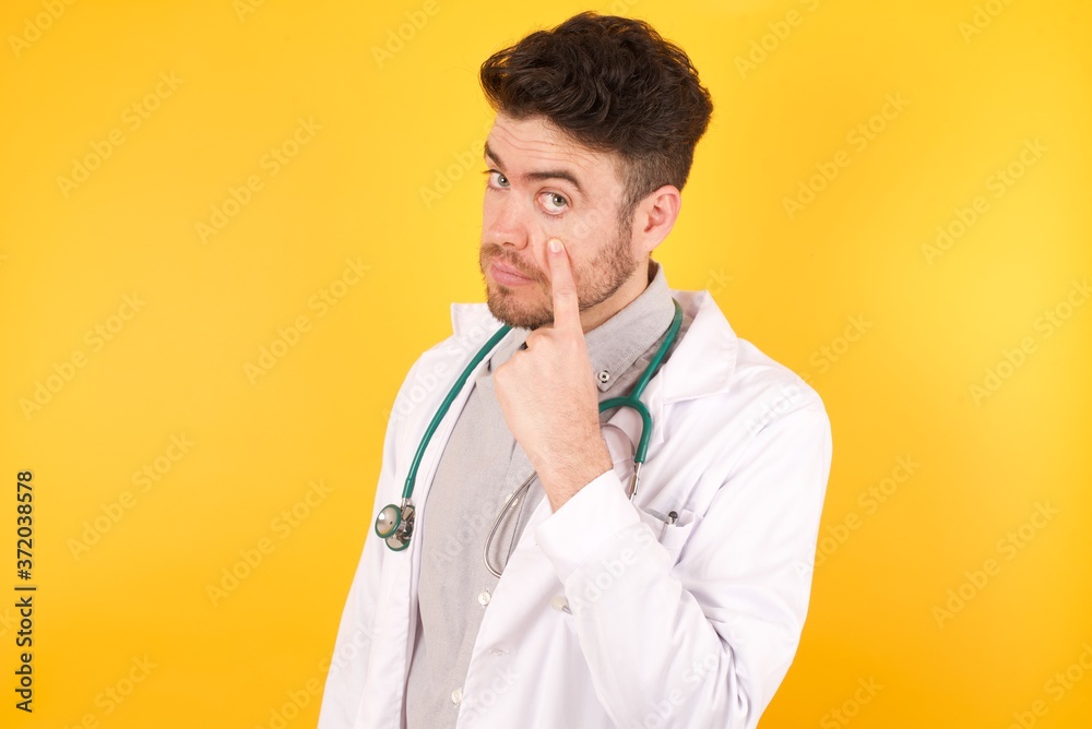 Young handsome Caucasian doctor man wearing medical uniform standing over yellow background Pointing to the eye watching you gesture, suspicious expression. Young handsome Caucasian doctor man wearing medical uniform standing over yellow background Pointing to the eye watching you gesture, suspicious expression.
