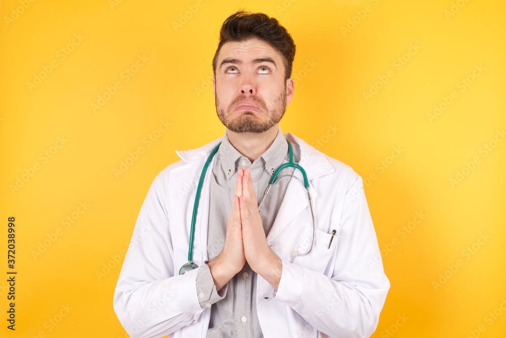 Young Caucasian doctor man wearing medical uniform over isolated yellow background begging and praying with hands together with hope expression on face very emotional and worried. Asking Young Caucasian doctor man wearing medical uniform over isolated yellow background begging and praying with hands together with hope expression on face very emotional and worried. Asking