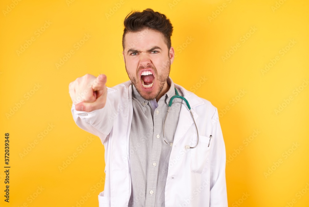 Young handsome Caucasian doctor man wearing medical uniform standing over isolated yellow background pointing displeased and frustrated to the camera, angry and furious ready to fight with you. Young handsome Caucasian doctor man wearing medical uniform standing over isolated yellow background pointing displeased and frustrated to the camera, angry and furious ready to fight with you.