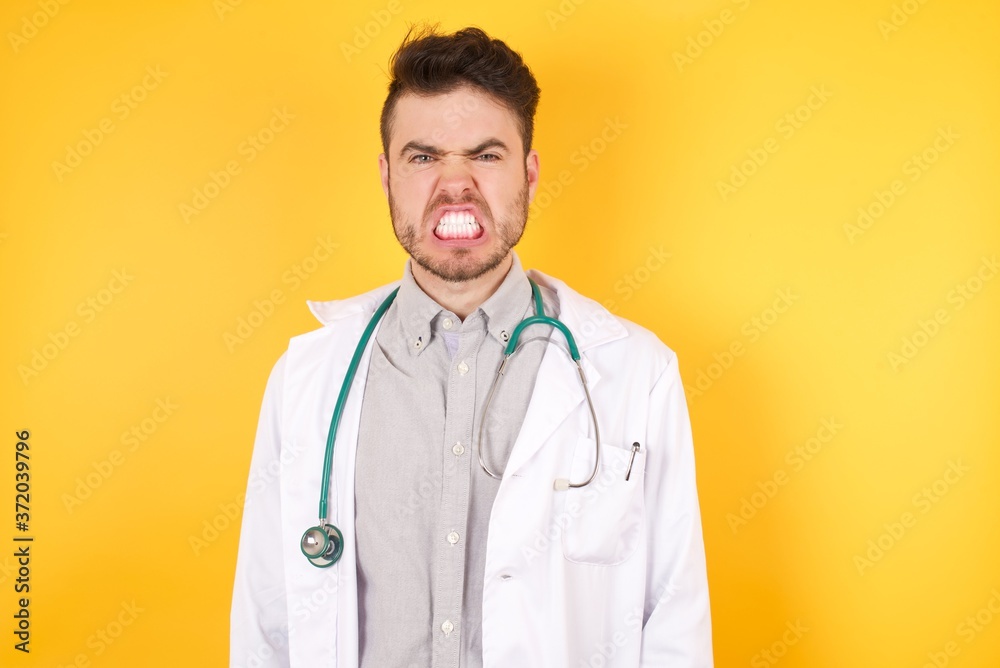 Portrait of angry doctor man wearing medical uniform, keeps teeth clenched, frowns face in dissatisfaction, irritated because of much duties. Furious man looks out of window, sees miserable weather Portrait of angry doctor man wearing medical uniform, keeps teeth clenched, frowns face in dissatisfaction, irritated because of much duties. Furious man looks out of window, sees miserable weather