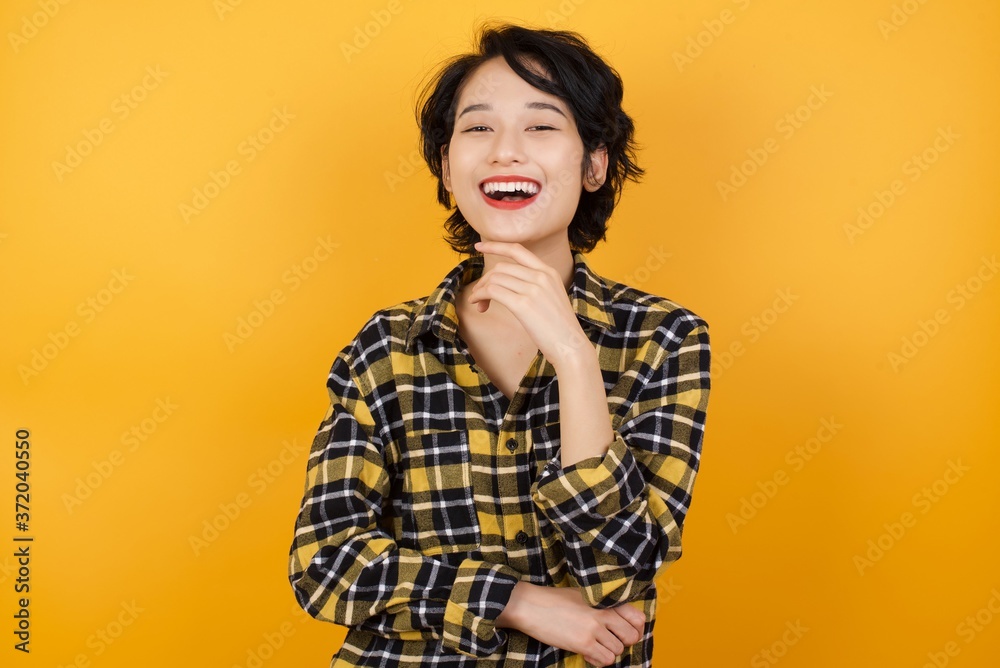 Optimistic  Young asian woman with short hair wearing plaid shirt standing over yellow background keeps hands partly crossed and hand under chin, looks at camera with pleasure, Happy emotions concept.