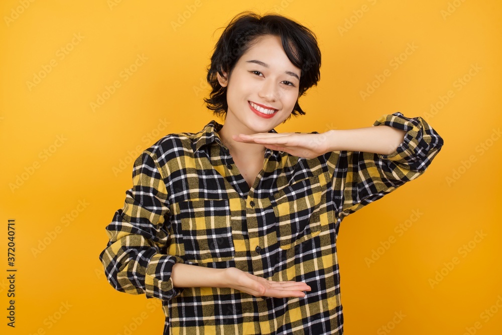Young asian woman with short hair wearing plaid shirt standing over yellow wall gesturing with hands showing big and large size sign, measure symbol. Smiling looking at the camera. Measuring concept. Young asian woman with short hair wearing plaid shirt standing over yellow wall gesturing with hands showing big and large size sign, measure symbol. Smiling looking at the camera. Measuring concept.
