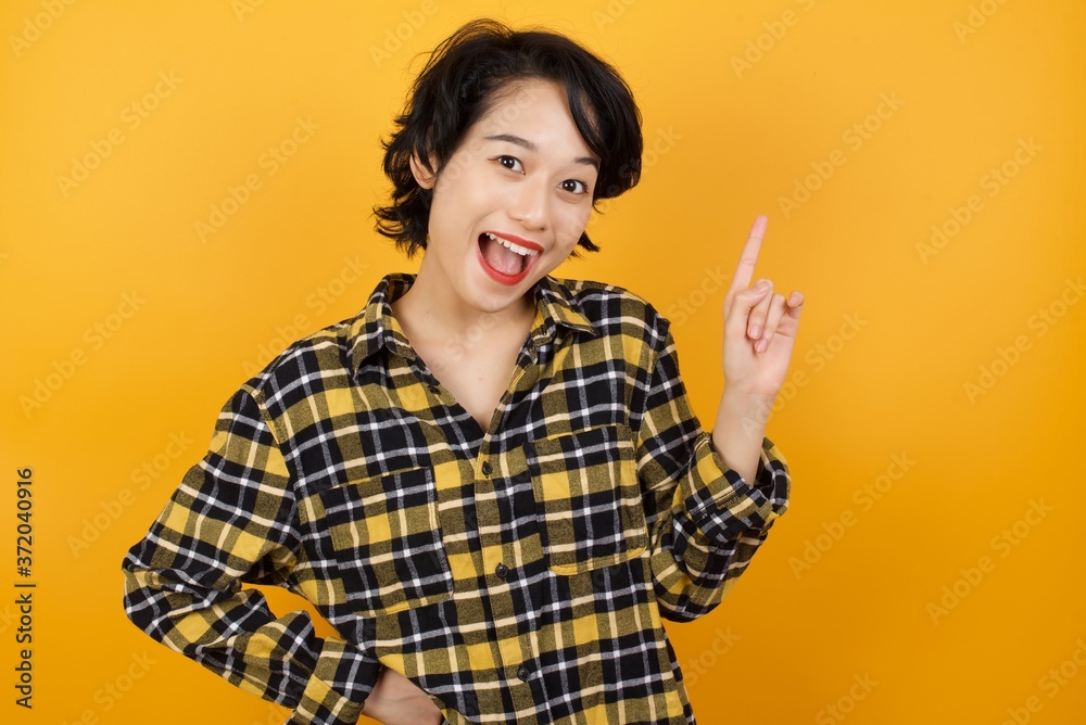 Young asian woman with short hair wearing plaid shirt standing over yellow background holding finger up having idea and posing Young asian woman with short hair wearing plaid shirt standing over yellow background holding finger up having idea and posing