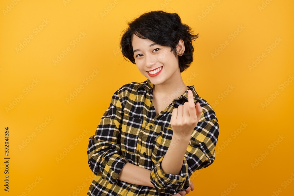 Young asian woman with short hair wearing plaid shirt standing over yellow background Beckoning come here gesture with hand inviting welcoming happy and smiling Young asian woman with short hair wearing plaid shirt standing over yellow background Beckoning come here gesture with hand inviting welcoming happy and smiling