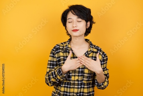 Young beautiful Asian woman wearing plaid shirt over yellow background smiling with hands on chest with closed eyes and grateful gesture on face. Health concept.