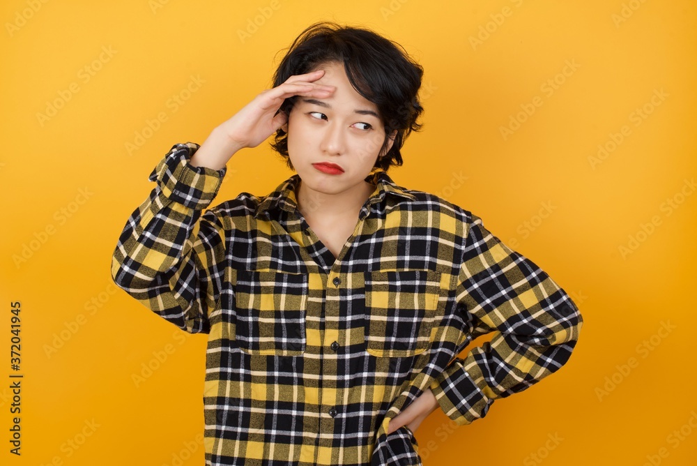 Young beautiful Asian woman wearing plaid shirt over yellow background having problems, worried and stressed holds hand on forehead.