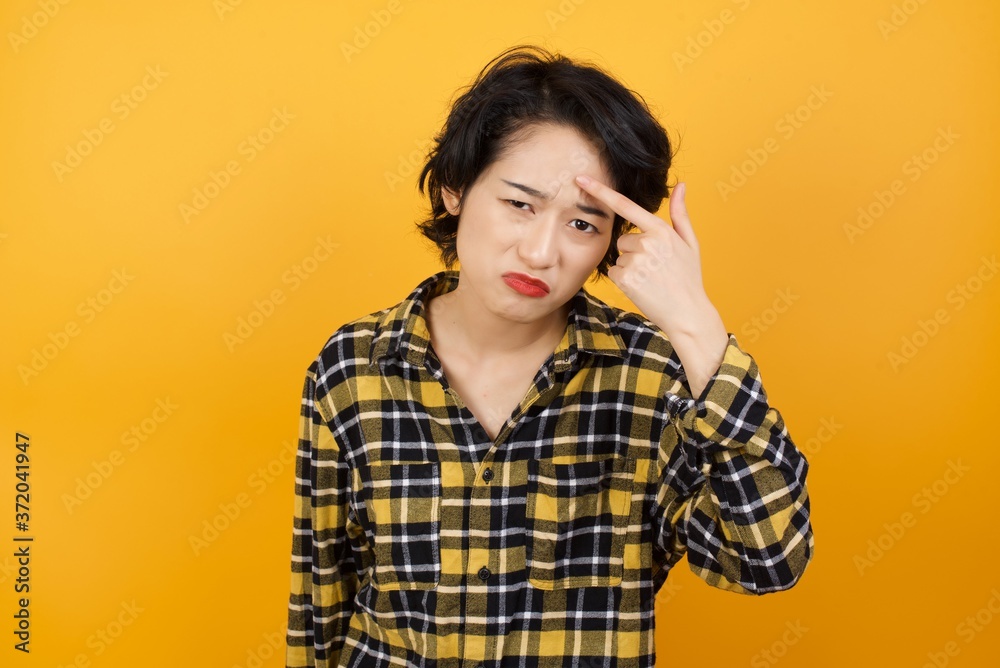 Young beautiful Asian woman wearing plaid shirt over yellow background pointing unhappy at pimple on forehead, blackhead infection. Skincare concept. Young beautiful Asian woman wearing plaid shirt over yellow background pointing unhappy at pimple on forehead, blackhead infection. Skincare concept.