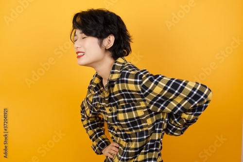 Young beautiful Asian woman wearing plaid shirt over yellow background Suffering of backache, touching back with hand, muscular pain
