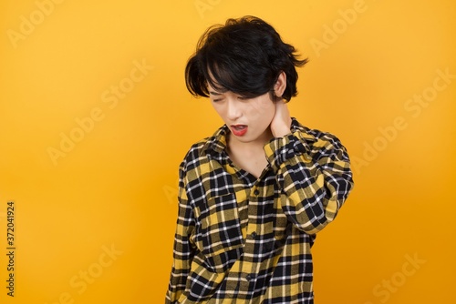 Young beautiful Asian woman wearing plaid shirt over yellow background Suffering of neck ache injury, touching neck with hand, muscular pain