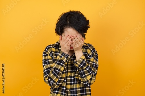 Young beautiful Asian woman wearing plaid shirt over yellow background covering her face with her hands, being devastated and crying.