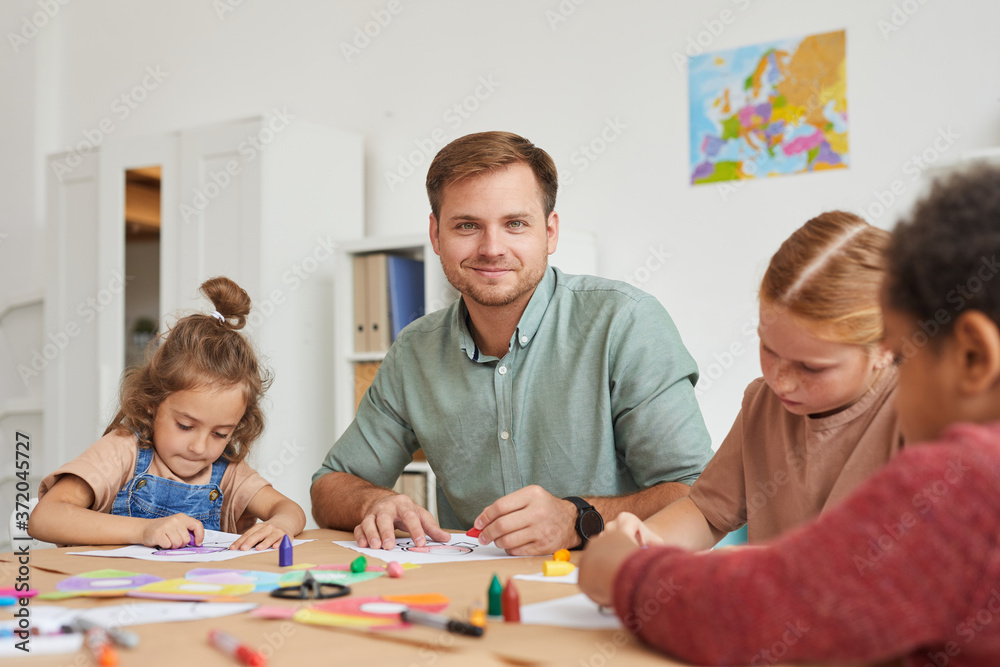 Obraz premium Portrait of male teacher smiling at camera while working with multi-ethnic group of children drawing pictures during art class in school or development center, copy space