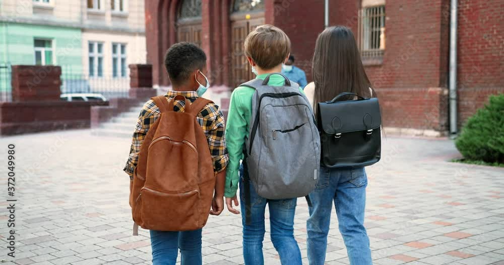 Rear of multi-ethnic junior students with backpacks going to school. Caucasian girl and boy. African American pupil with schoolbag. Male and female teachers on background tapping on smartphone