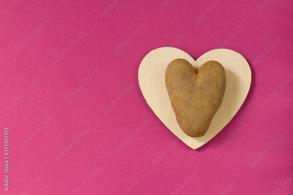 Obraz premium Ugly potatoes in the shape of a heart on a wooden board on a pink background. Unusual concept of vegetables or food waste. horizontal orientation. Top view. Copy space