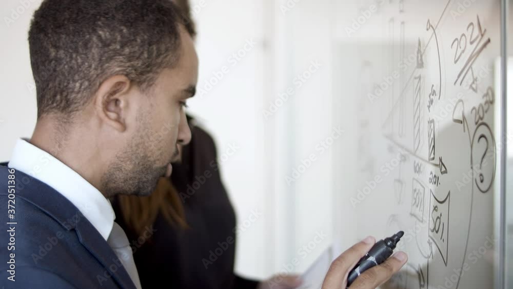 Serious business man drawing chart on glass board. Colleagues analyzing paper report. Closeup shot, side view. Analysis and teamwork concept Serious business man drawing chart on glass board. Colleagues analyzing paper report. Closeup shot, side view. Analysis and teamwork concept