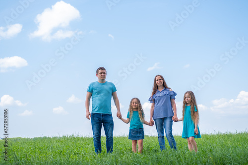 family dad, mom and two children walking on green grass against blue sky. Happy caucasian parents with two daughters holding hands.
