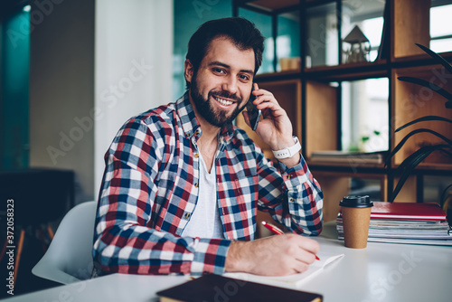 Portrait of happy handsome hipster guy posing and looking at camera while making conversation via telephone, positive male student learning and using smartphone for call indoors during coffee break