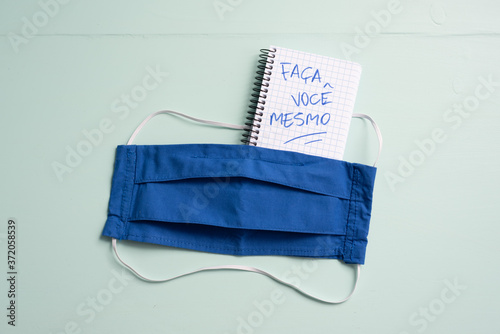 Notepad written Faça Você Mesmo -Do It Yourself- in portuguese and crafted facemask flat lay with copy space of aqua menthe desk photo