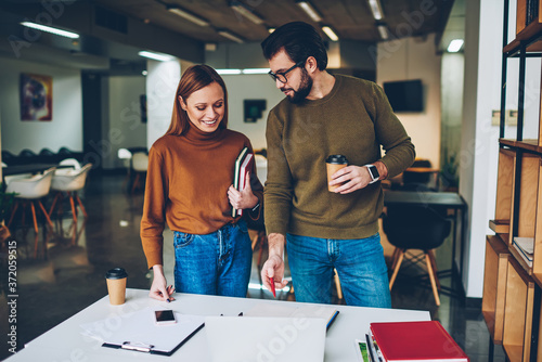 Two young casually dressed colleagues standing together in large modern office having coffee break from tired paperwork, successful hipsters working while male leader explaining information indoors