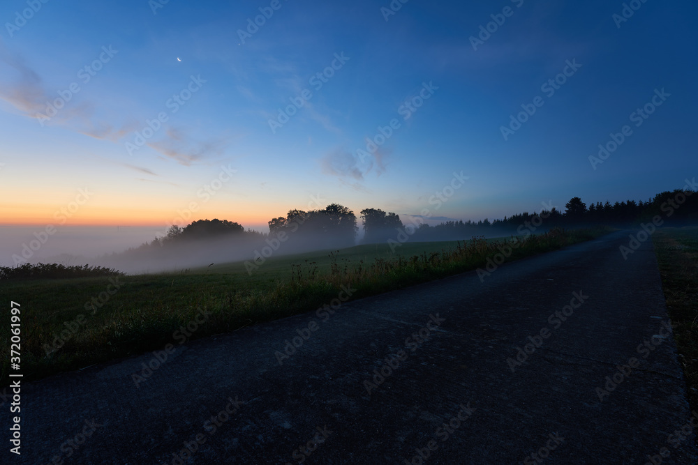 Sunrise in August, celestial bodies moon and Venus in the blue sky, construction crane can be seen in the fog in the distance, dark road in the foreground Germany, Swabian Alb.