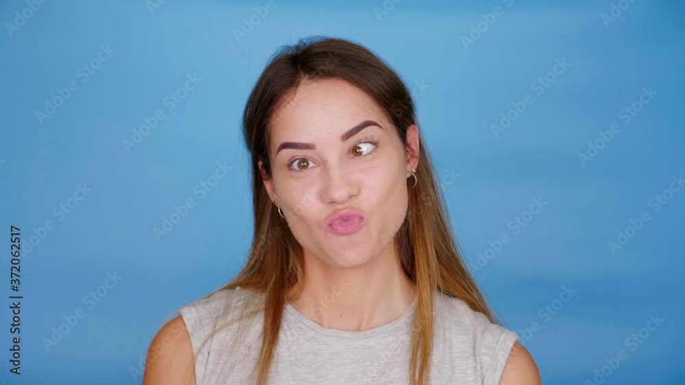 Beautiful funny woman in gray t-shirt squints and smiles on blue background with copy space. 4K slow motion footage Beautiful funny woman in gray t-shirt squints and smiles on blue background with copy space. 4K slow motion footage