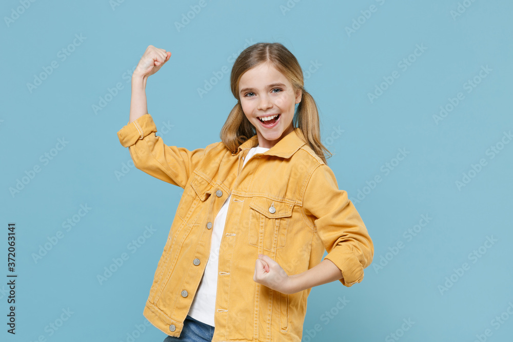 Happy little blonde kid girl 12-13 years old in yellow jacket posing isolated on blue background children studio portrait. Childhood lifestyle concept. Mock up copy space. Clenching fists like winner. Happy little blonde kid girl 12-13 years old in yellow jacket posing isolated on blue background children studio portrait. Childhood lifestyle concept. Mock up copy space. Clenching fists like winner.